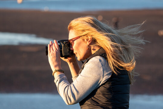A woman with a DSLR takes an outdoor picture on a windy day. Her hair blows in the wind as she focuses the camera. Warm light at sunset casts across her.
