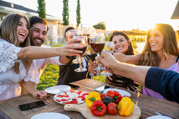 Group of friends celebrating in the garden in front of the house toasting a table set with glasses of red wine - Young people having fun together laughing and joking on a summer evening - Copy space