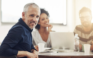 Im proud to lead this team. Portrait of a mature office worker sitting at a table with colleagues in the background.