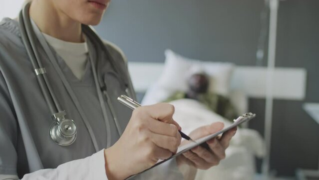 Tilt up medium close-up of Caucasian female practitioner standing military hospital room filling in patients medical card