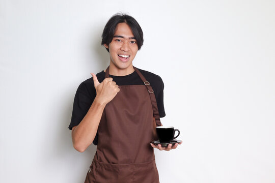 Portrait of attractive Asian barista man in brown apron with thumb pointing away on empty space. Advertising concept. Isolated image on white background