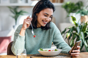 Beautiful young woman eating a bowl of salad while usingh her mobile phone in the living room at...