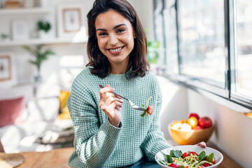 Beautiful smiling woman eating healthy salad while sitting on the table at home. - obrazy, fototapety, plakaty