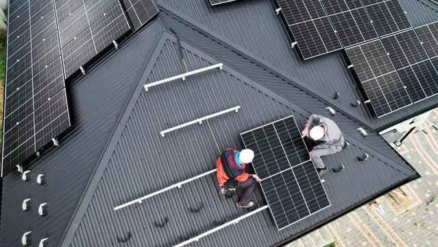 Technicians installing photovoltaic solar panels on roof of house. Drone view of men engineers in helmets building solar module system with help of hex key. Concept of alternative, renewable energy.