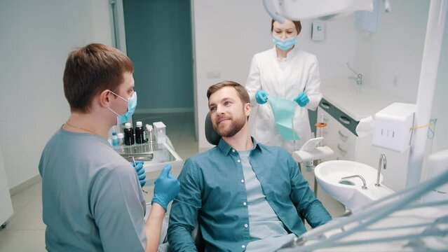A male doctor discusses a dental treatment plan with a patient. Dentistry. Dental clinic. In the background, a female doctor is preparing a patient