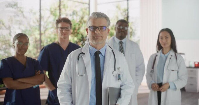 Group of Successful Multiethnic Team of Female and Male Doctors, Nurses and Healthcare Professionals Posing for Camera and Smiling. Portrait of a Confident Middle Aged Physician Standing First in Line