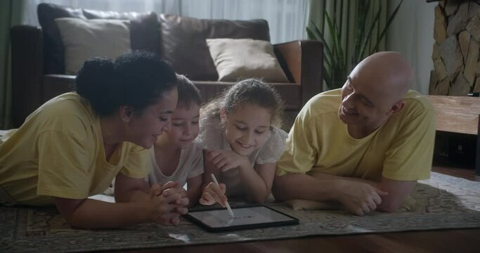 Happy family young parents drawing coloring picture with pencils helping cute child daughter enjoying talk play laying on warm floor at home, mom dad and kid girl having fun in living room on leisure