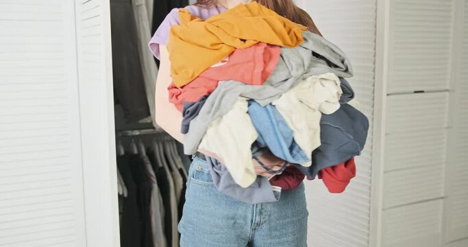 Woman with lot of clothes in her hands turns back and forth in front of camera. Concept of abundance of things, washing, happiness, satisfaction, shopaholic.