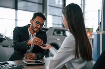 Woman is consulting man in the car showroom. Taking keys from the new automobile