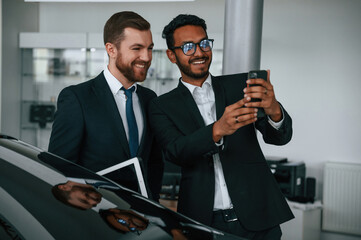Fototapeta na wymiar Having fun, using smartphone. Two businessmen are working together in the car showroom