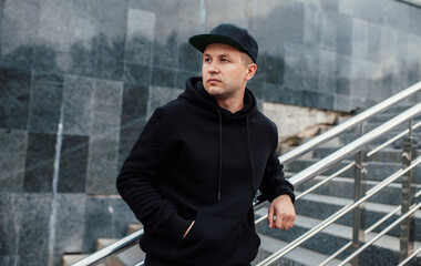 City portrait of handsome guy wearing black blank hoodie or sweatshirt and baseball cap with space...