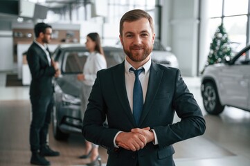 Fototapeta premium Beautiful man is standing and smiling. Three people are working together in the car showroom