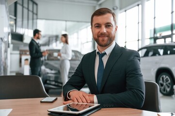 Man in headphones is sitting by table. Three people are working together in the car showroom