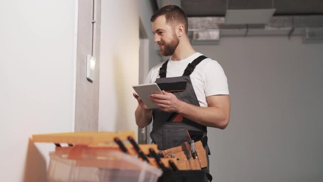 Portrait of a man in a company work uniform with a tool belt and tablet in hand, configuring an access panel for controlling ventilation, air conditioning, security alarm, and surveillance cameras
