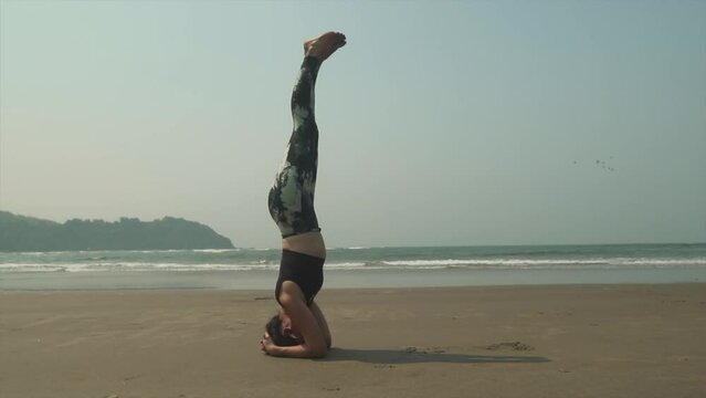 Fit young  woman practicing a yoga head stand, Salamba Shirshasana, on a warm tropical beach. 