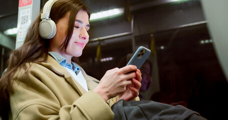 Low angle side view of cute beautiful young woman travelling by bus listening to music chatting online with smartphone. Pretty caucasian female smiling enjoying ride in empty public transport.