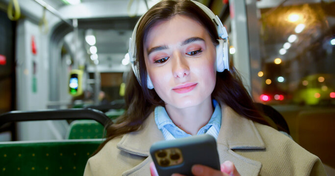 Close-up portrait of charming calm young woman listening to music in wireless headphones using smartphone sitting in bus. Beautiful joyful caucasian female using public transport smiling.