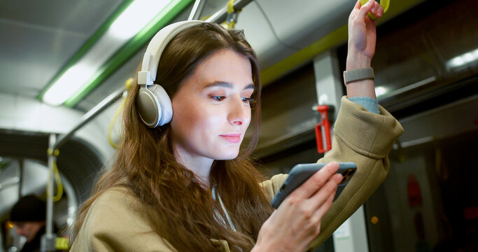 Side-view Portrait Of Focused Cute Woman Watching Video On Smartphone Using Headphones Riding Bus. Attractive Happy Caucasian Female Standing In Public Transport Holding Handrail.