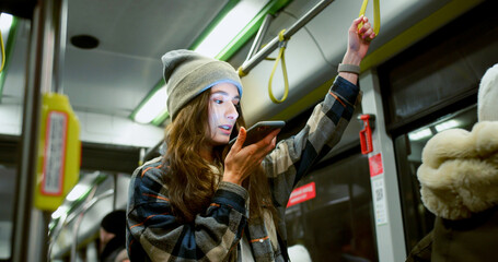 Stylish young woman standing in bus recording and listening to voice messages holding handrail. Attractive caucasian adult girl using smartphone wearing shirt and beanie. Public transport concept.