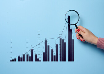 A woman's hand holds a magnifying glass and a graph with growing indicators, studying data,...