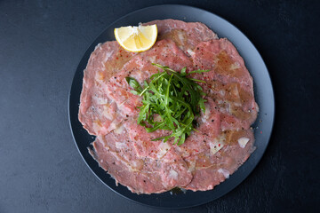 Marbled beef carpaccio with arugula, parmesan cheese, olive oil and lemon. A traditional classic appetizer made from thinly sliced raw meat. Close-up, selective focus, top view, black background.