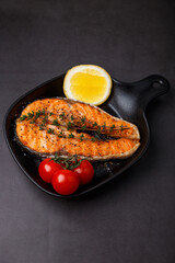 Grilled salmon with cherry tomatoes, thyme and lemon on a small black grill pan. Fried fish is a traditional dish of home cooking and restaurants. Black background, selective focus, close-up.