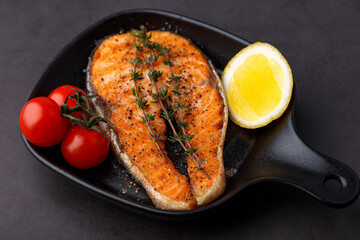 Grilled salmon with cherry tomatoes, thyme and lemon on a small black grill pan. Fried fish is a traditional dish of home cooking and restaurants. Black background, selective focus, close-up.