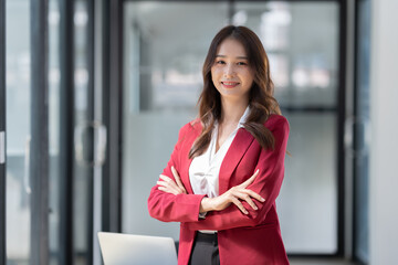 Asian businesswoman or freelancer standing near the desk in the office Dressed in formal clothes, crossed arms, looking at the camera, smiling friendly.