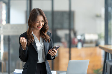 Asian businesswoman holding mobile phone with laptop looking at information laughing at news for online shopping happy.