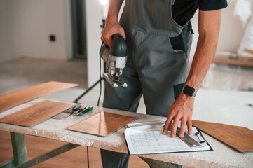 Standing by the table with jigsaw. Man is installing new laminated wooden floor