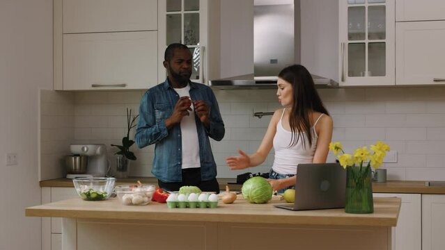 African american black man and caucasian woman cooking together in the kitchen at home. The male is juggling tomatoes while the femele asks him not to fool around.