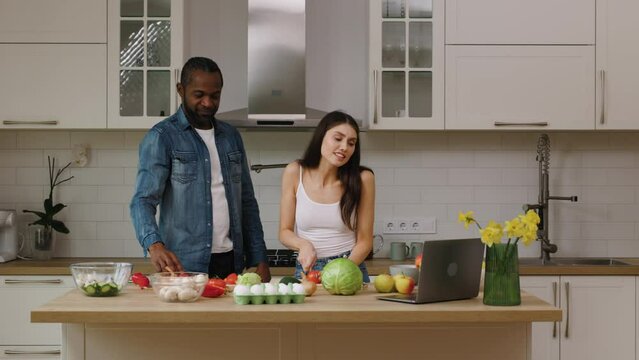 African american man and caucasian woman are making salad together in the kitchen at home. The couple is talking with friends via videocall.