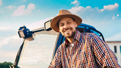 Close up of Caucasian handsome young male farmer in hat at tractor, smiling cheerfully and looking at camera. Farming concept. Portrait of attractive man having rest from work at farm. Outdoor.