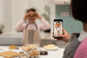 Happy european adult woman taking photo on smartphone screen of little girl in apron, making cookie - obrazy, fototapety, plakaty