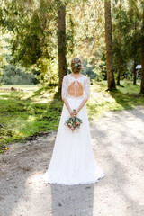 Fototapeta premium Braut im Brautkleid mit ausgeschnittenem Rücken hält Brautstrauß bei Hochzeit im Wald