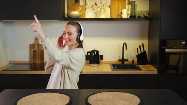Young amazing emotional woman dancing in the kitchen indoors at home, listening to music with headphones. 