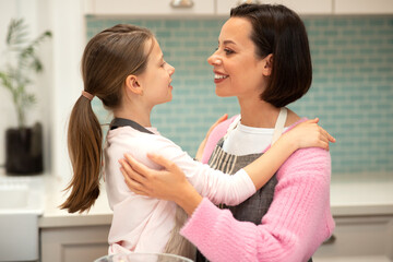 Smiling european millennial woman hugs little girl cheef in apron in kitchen interior, close up, profile