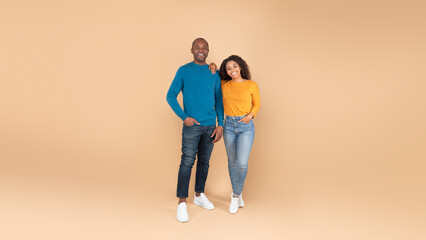Full body length portrait of happy black couple looking at camera and posing standing over peach background, panorama