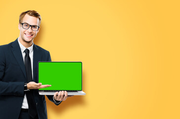 Business man in glasses spectacles, black suit show laptop with empty mockup green chroma key screen, isolated orange yellow background. It expert,  technician repair service, tech support maintenance