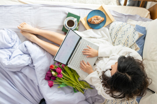 A young asian girl sits on the bed in the morning, uses a laptop, eats croissants and drinks coffee for breakfast. Enlarging a picture from the camera from top to bottom