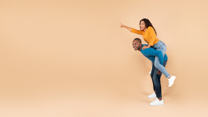 Fototapeta na wymiar Happy black woman piggybacking her husband and pointing at free space, peach studio background, copy space, panorama