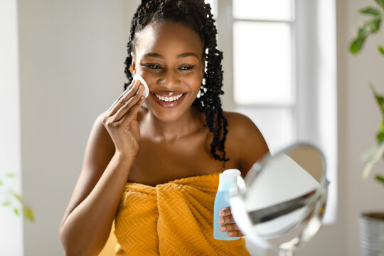 Face care routine. Young african american lady wrapped in towel holding bottle with micellar water and using cotton pad