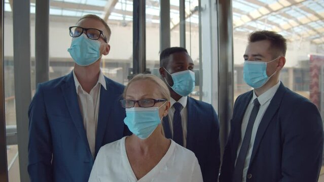 Four multiethnic businesspeople wearing protective face masks ride a glass elevator to office 