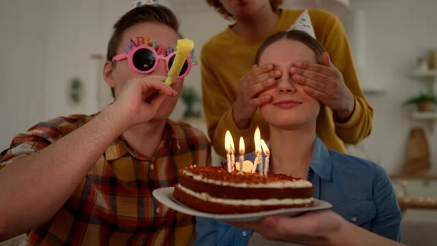 Surprise for birthday girl from friends. Friend closes eyes with hands. She happy about birthday cake with burning candles. Guy in funny glasses blowing party horn. Celebration, festive mood concept.