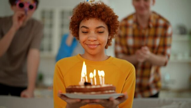 Portrait smiling african american birthday girl with cake in hands looks at burning candles makes a wish. Happy party with friends clapping hands and blowing horns on background. Celebration concept.
