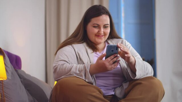 Plus size woman in bedroom chatting using smartphone. Realtime