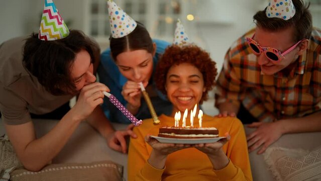 African american girl celebrates birthday with friends blows candles on cake makes a wish. They smile and clap hands. Wearing festive caps and blows party horns. Celebration party for birthday girl.