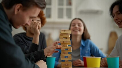 Friends playing leaning tower board game at home. Friends take turns pulling out wooden blocks and putting them up. Guy thinks, examines and builds strategy to win. Nervous game of skill and logic.