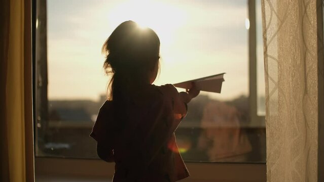 A child plays with a paper plane near the window of the house during sunset.Authentic scene.