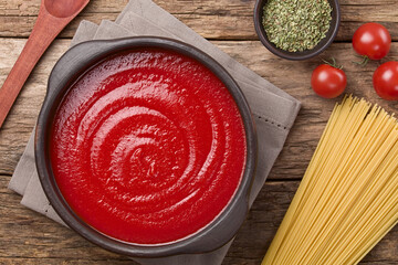 Tomato paste in rustic bowl, with dry spaghetti, cherry tomato, dried oregano and wooden spoon on the side, photographed overhead on wood